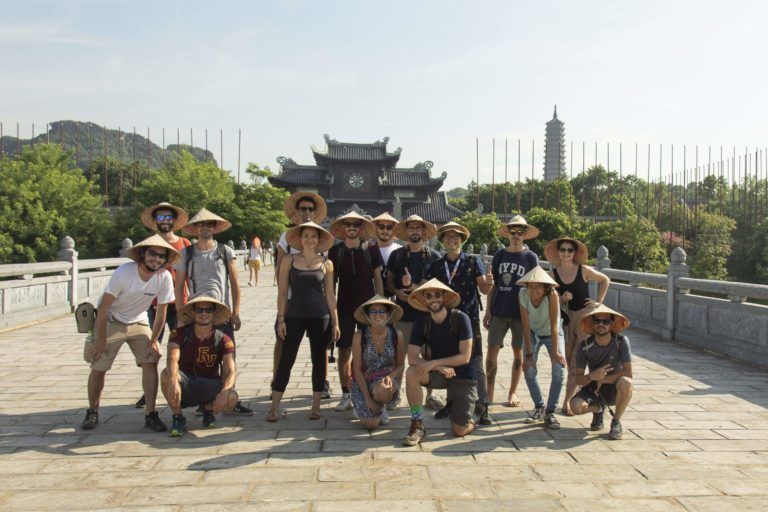 foto de grupo de viajeros de weroad con sombrero típico de vietnam en hue, detrás un templo y vegetación
