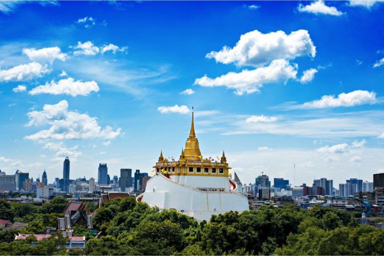 templo de wat saket, lugar que ver en bangkok, visto desde lejos. a sus pies vegetación, al fondo edificios modernos y cielo azul con nubes blancas - weroad