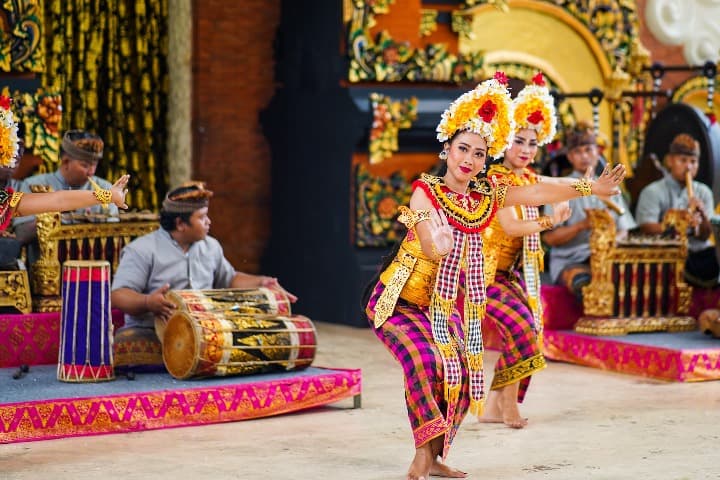 dos bailarinas en traje típico bailan una danza balinesa, detrás unos hombres sentados por el suelo tocacn varios instrumentos - weroad