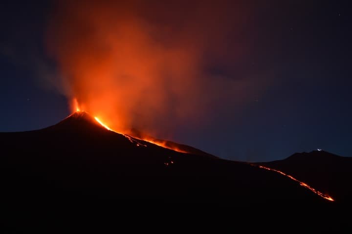 volcan etna eruptando de noche, humo y lava naranja con cielo de fondo - weroad