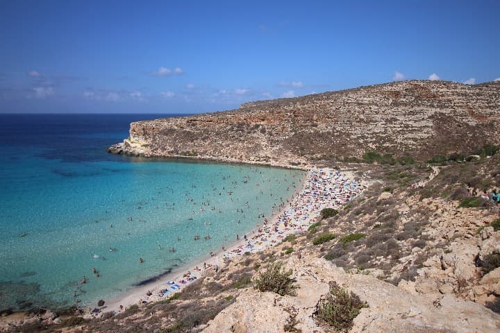playa de  isla dei conigli en lampedusa, mar turquesa con gente bañandose y al fondo montaña - weroad