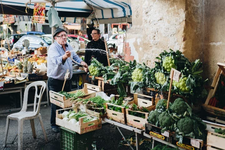 señor regando una parada de verduras verdes en el mercado de palermo, una de las cosas que ver en sicilia - weroad