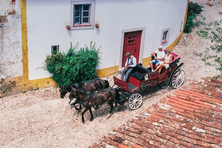 carro tirado por dos caballos y tres personas montadas en él por las calles de óbidos, ciudad que ver en portugal - weroad