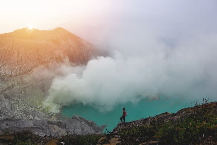 hombre visto desde lejos mientra observa el humo salir del volcan kawah ijen, una de las cosas que ver en indonesia, al fondo montañas, al lado agua azul - weroad