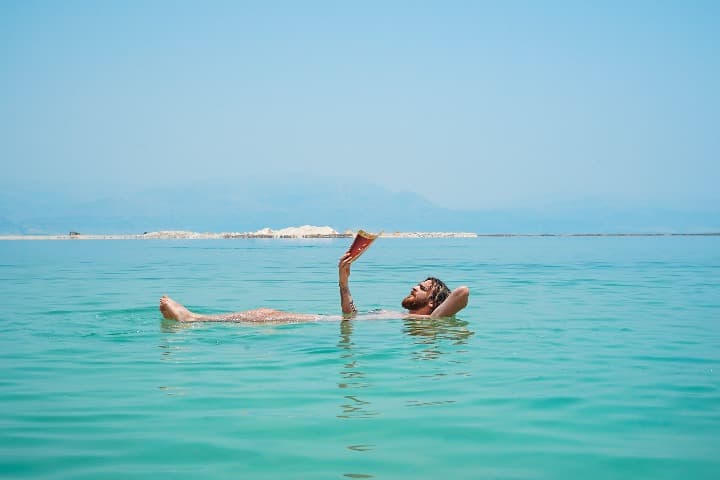 chico flotando en el mar muerto mientras lee un libro, agua verde, cielo azul - weroad