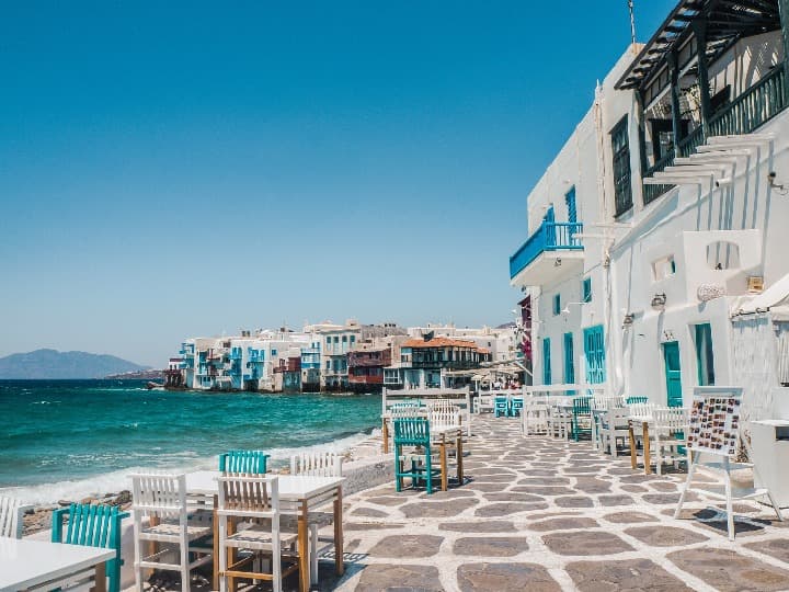 mesas y sillas en una calle de mykonos, grecia, a orillas del mar, al otro lado casas blancas con decoraciones azules y verdes. dónde ir de vacaciones en agosto - weroad
