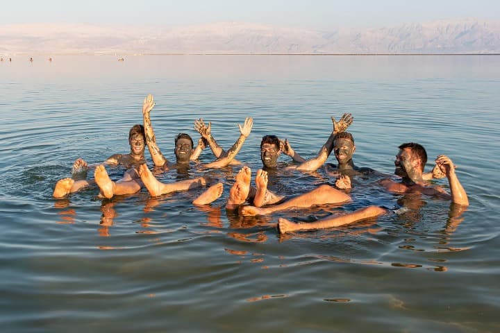 cinco chicos floatando en el mar muerto saludando cámara y con los rostros embadurnados de barro - weroad