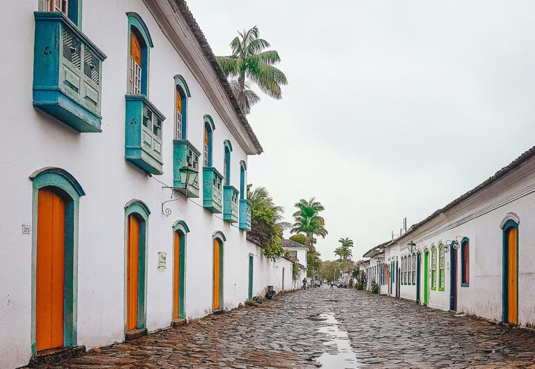 casas coloniales blancas y bajas con puertas naranja en una calle adoquinada en paraty, brasil. palmeras al fondo - weroad