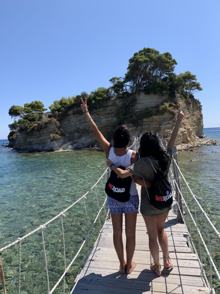 dos viajeras con mochila weroad de espaldas en un puente de madera suspendido en el agua mirando una roca en la isla cameo, zante