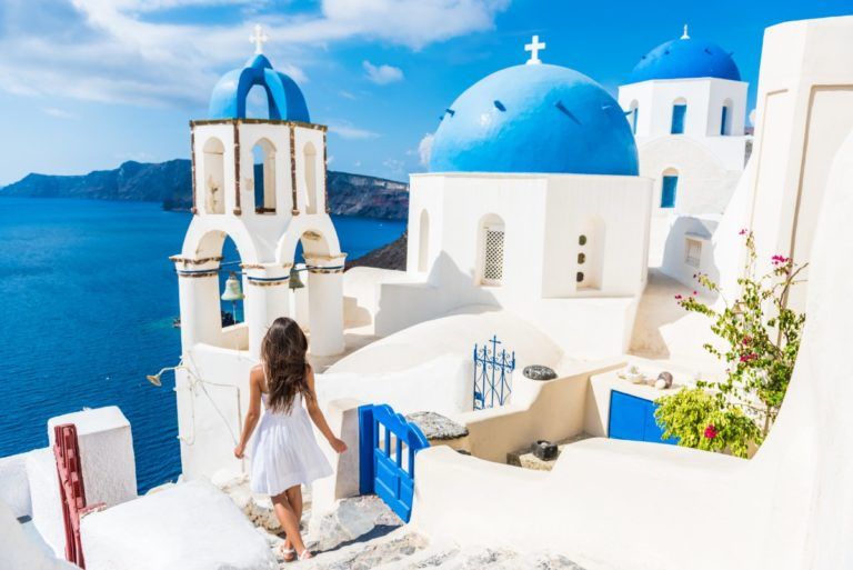 chica con vestido blanco bajando escaleras por las calles de santorini, una de las islas griegas más famosas. Edificios blancos con cúpolas aules. Al fondo el mar y el cielo azules - weroad