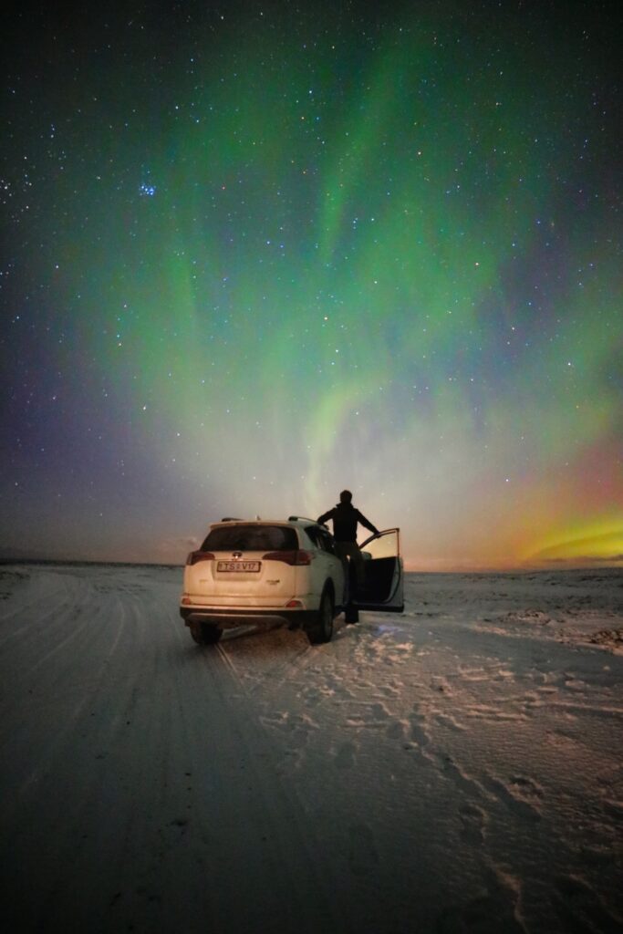 la aurora boreal es algo que ver en islandia, aurora boreal. persona de espaldas asomandose fuera de un coche blanco en medio de la nieve mirando hacia el cielo de color verde, aurora boreal - weroad