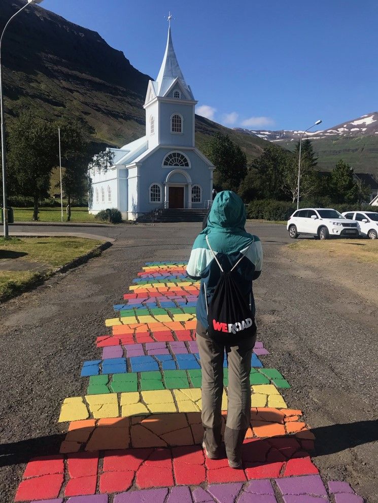 viajero de espaldas con mochila weroad en rainbow street en seyðisfjörður, en islandia. en el fondo una iglesia azul