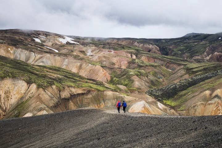 dos personas con mochila azul caminando por senderos en medio de montañas en landmannalaugar - weroad