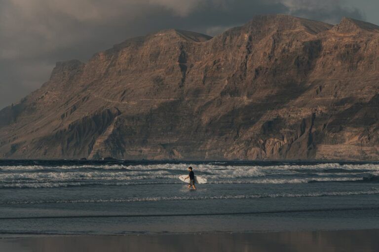 surfista en playa de lanzarote caminando por la orilla de playa famara llevando la tabla bajo el brazo, en el fondo una montaña