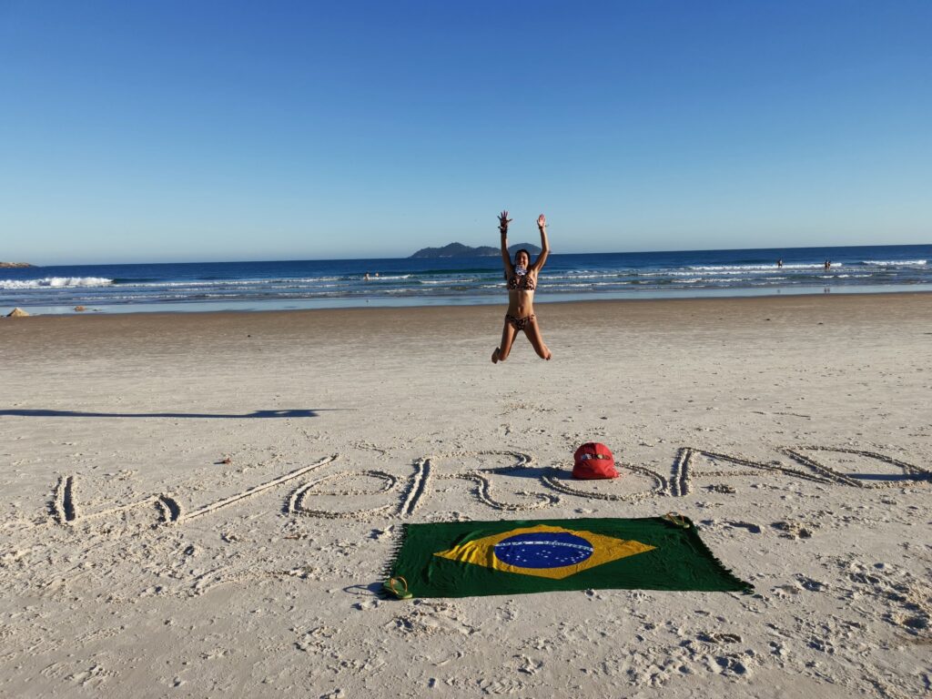 chica saltando en una playa de brasil con escrito weroad en la arena, al lado una bandera del país