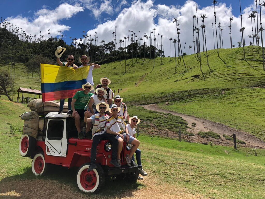 grupo de viajeros de weroad sentados encima de un coche en salento, aguantan una bandera de colombia, detrás colinas verdes