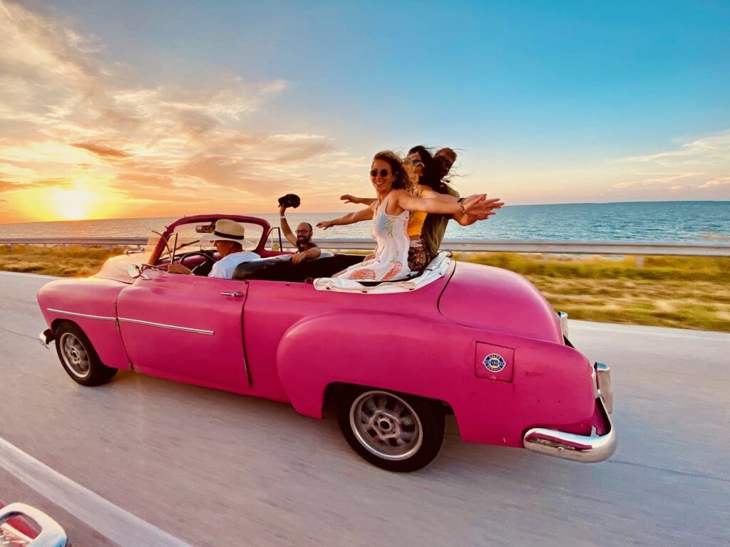coche descapotable de época color rosa chicle con tres chicos sentados en el asiento de atrás saludando a cámara, detrás mar con puesta del sol, cuba - weroad