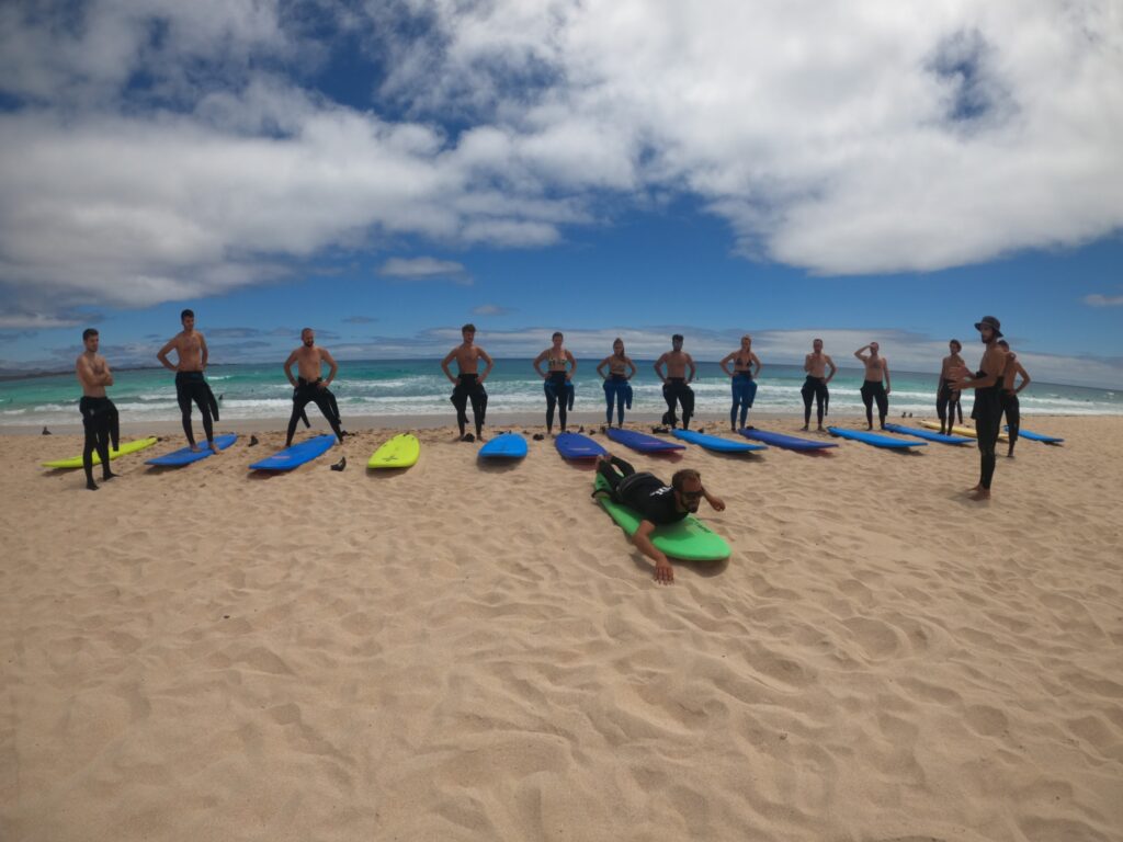 13 viajeros de weroad haciendo una clase de surf en la playa, en fila, cada uno con una tabla de surf delante suyo en la arena, uno imita los movimientos encima de su tabla al suelo, detrás el mar