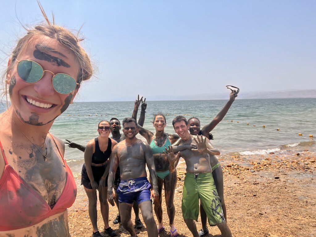selfie de viajera weroad con detrás grupo de compañeros embadurnados de barro en el mar muerto en jordania