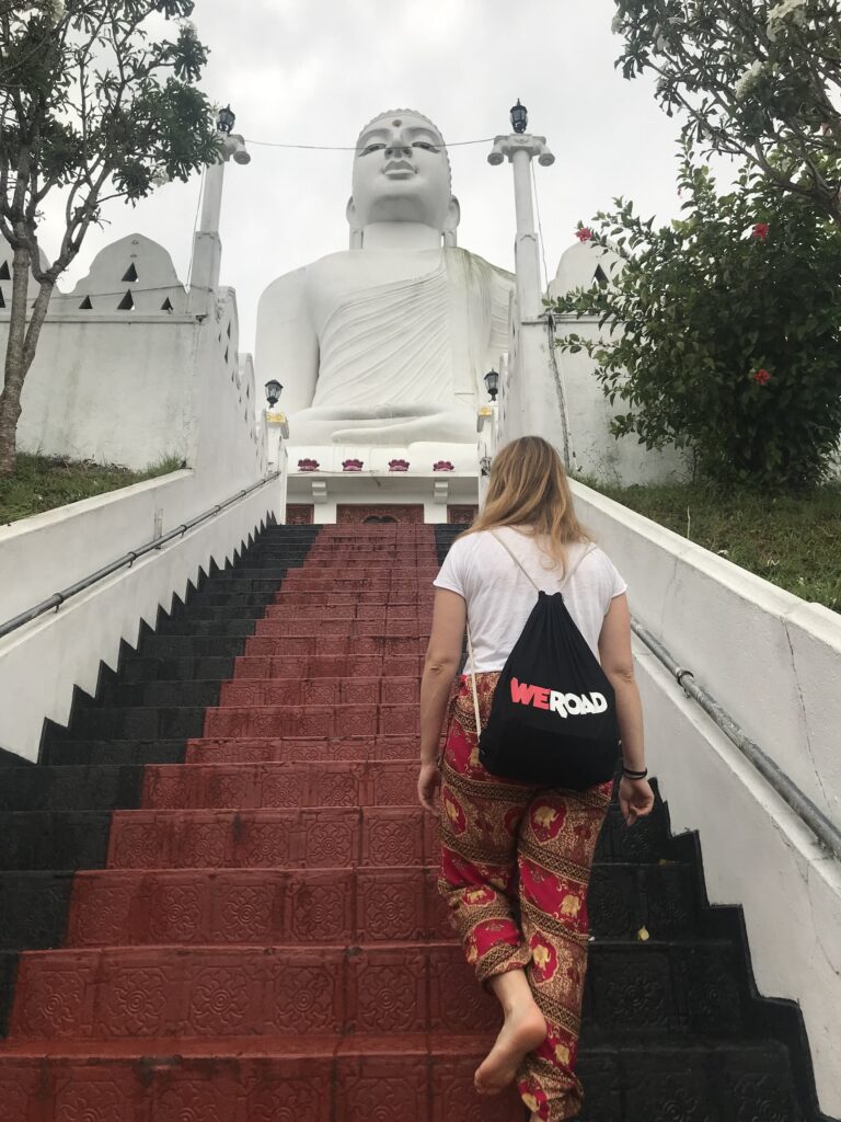 chica con mochila de weroad sube descalza unas escaleras rojas dirigiéndose hacia una enorme estatua blanca de buda