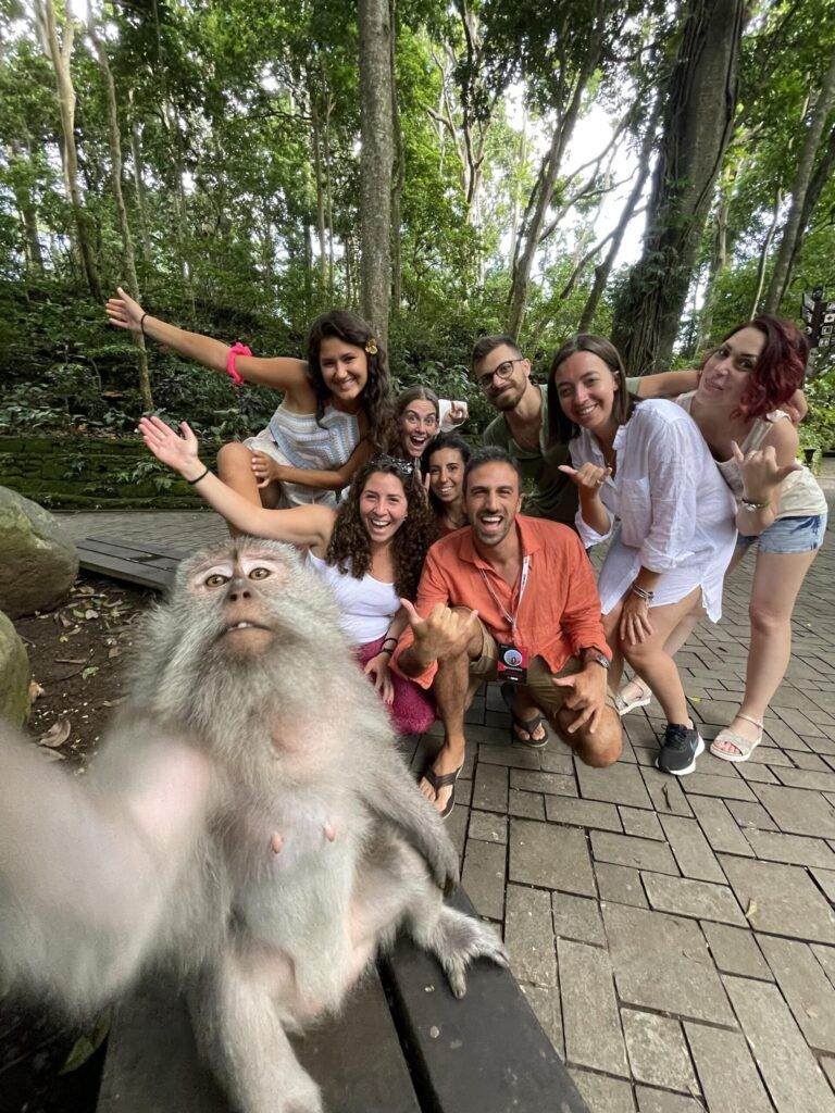 Selfie divertido de un grupo de viajeros de WeRoad posando con un mono de Bali, en un entorno de selva. El mono está en primer plano mirando a la cámara.