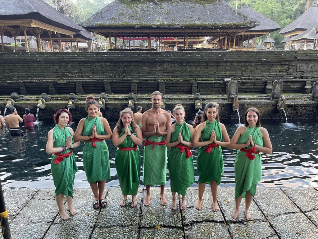 siete viajeros de weroad mirando a cámara con detrás fuentes del tirta empul, templo que ver en bali,. van vestidos con traje verde y cinturón rojo y tienen las manos cruzadas