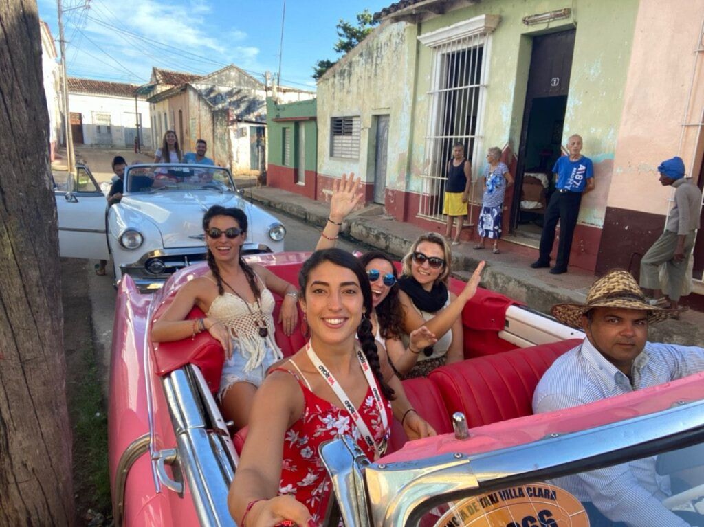 cuatro viajeros weroad haciedno un selfie en un coche antiguo en cuba, detrás otro coche antiguo con más personas