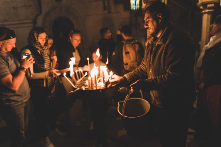 hombre con cubo que toca velas en el interior de la basílica del santo sepulcro, algo que ver en jerusalén. Al fondo más personas en la penumbra - weroad