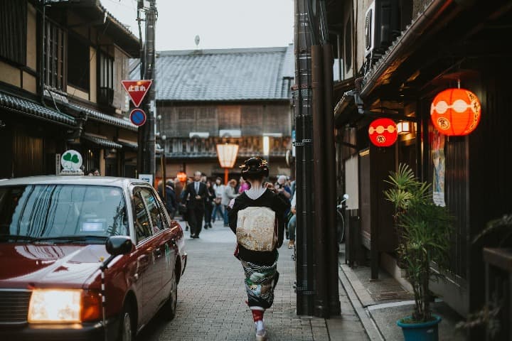 chica vestida de forma tradicional japonesa de espaldas caminando por el barrio de higashiyama, a su izquierda un coche, al fondo casas al estilo de montaña - weroad