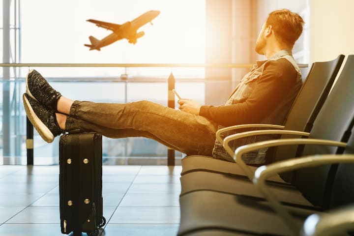 chico en el aeropuerto sentado y con pies encima de equipaje de mano, detras avion despegando - weroad