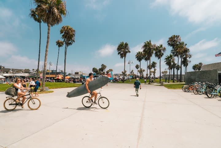 personas en bici, una lleva una tabla de surf, detrás palmeras y cesped en venice beach, california - weroad