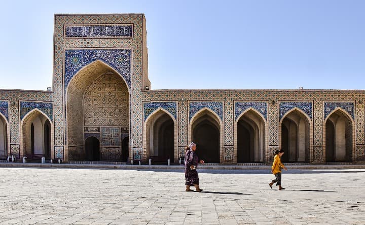 dos personas paseando delante de un edificio con arcos en bukkhara, ciudad que ver en uzbekistan
