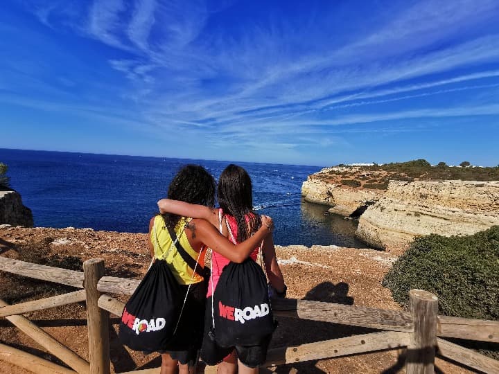 dos chicas de espaldas, abrazadas, con mochila de weroad, mirando el mar de algarve y un acantilado