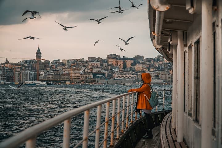 mujer con impermeable naranja encima de un barco, al fondo edificios de la ciudad de estambul y encima pajaros volando - weroad