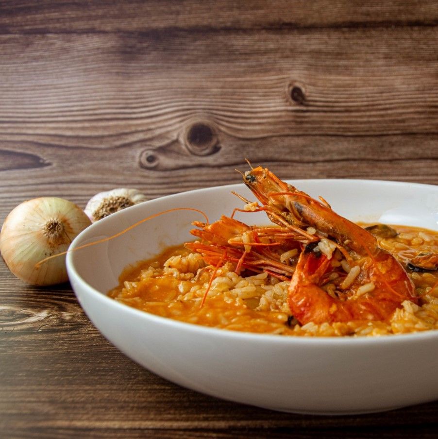 plato hondo encima de mesa de madera con en su interior arroz caldoso con marisco y un bogavante en primer plano, detrás cebolla y ajo. - weroad