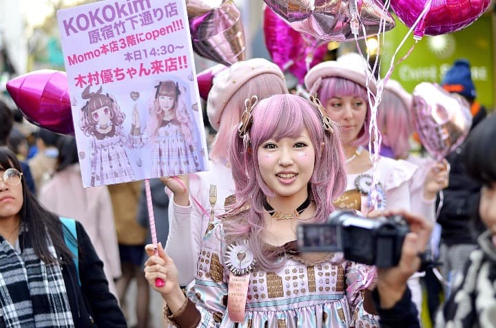 chica japonesa vestida de rosa con pelo rosa y aguantando un cartel con caracteres japoneses, en medio de más gente - weroad