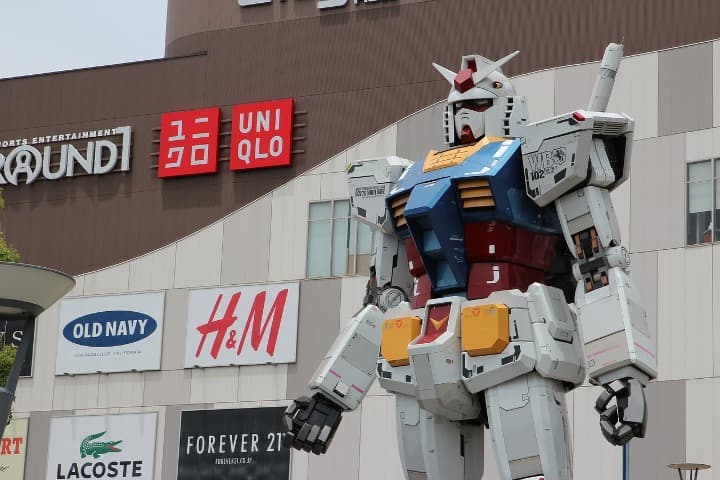 estatua de un Gundam Unicorn RX-0 delante de un centro comercial, en la fachada del edificio detrás aparecen logos de marcas de ropa - weroad