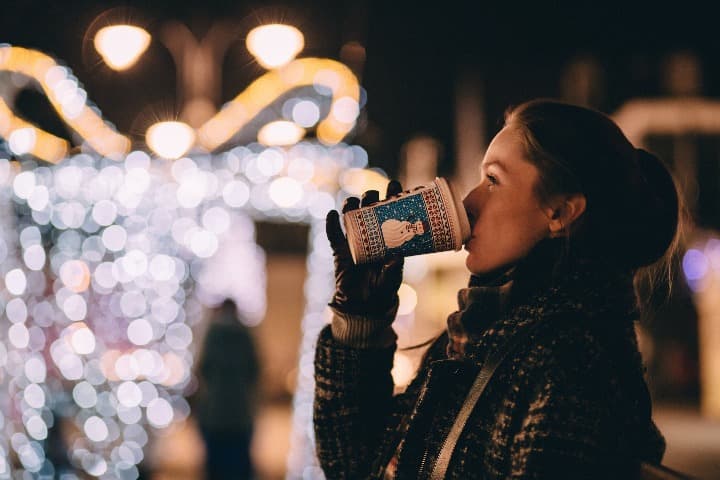 chica bebiendo de un vaso con detrás luces en un mercadillo de navidad en europa