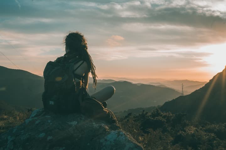 chica de espaldas ante una puesta del sol sentada encima de ujna piedra en medio de la montaña