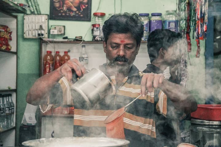 vendedor callejero de chai, en la india, haciendo te en su tienda, tiene un colador en una mano y un cazo en la otra, humo