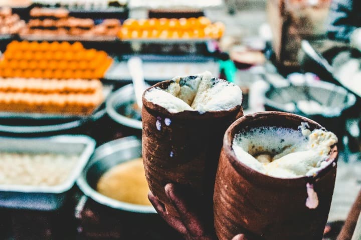 vasos de terracota con dentro lassi, detrás se ven pastelitos expuestos en una tienda, comida típica india