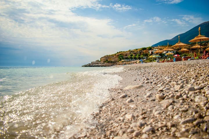 Dhërmi, una de las playas de albania más bonitas, foto del agua y del arena, sombrillas al fondo