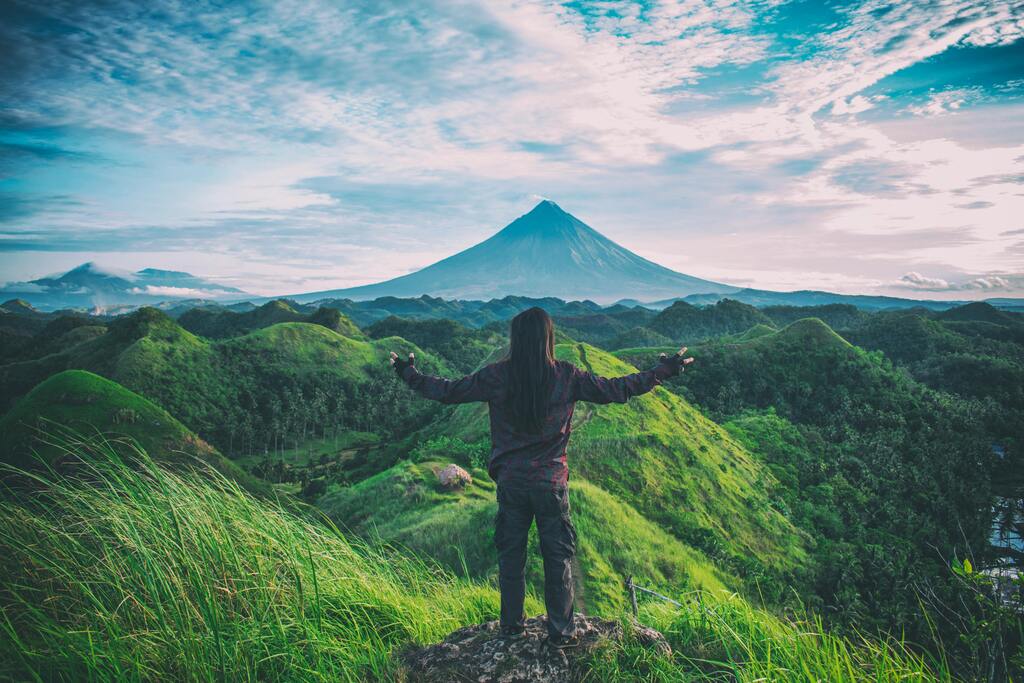 Viajero en la cima de una colina verde admirando el volcán Mayon en Filipinas, con un paisaje natural impresionante.