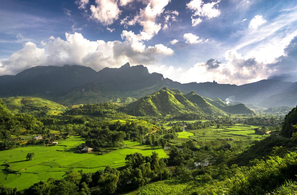 Vista de un valle verde en Vietnam con montañas al fondo bajo un cielo parcialmente nublado.
