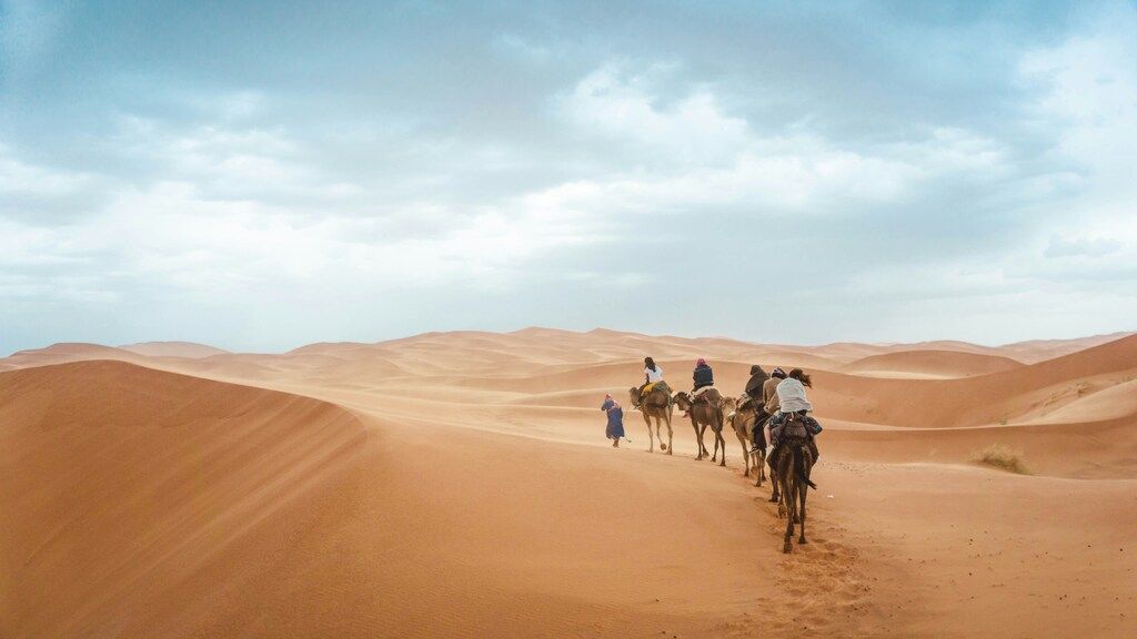 Grupo de viajeros montando camellos a través de las dunas del desierto bajo un cielo nublado.