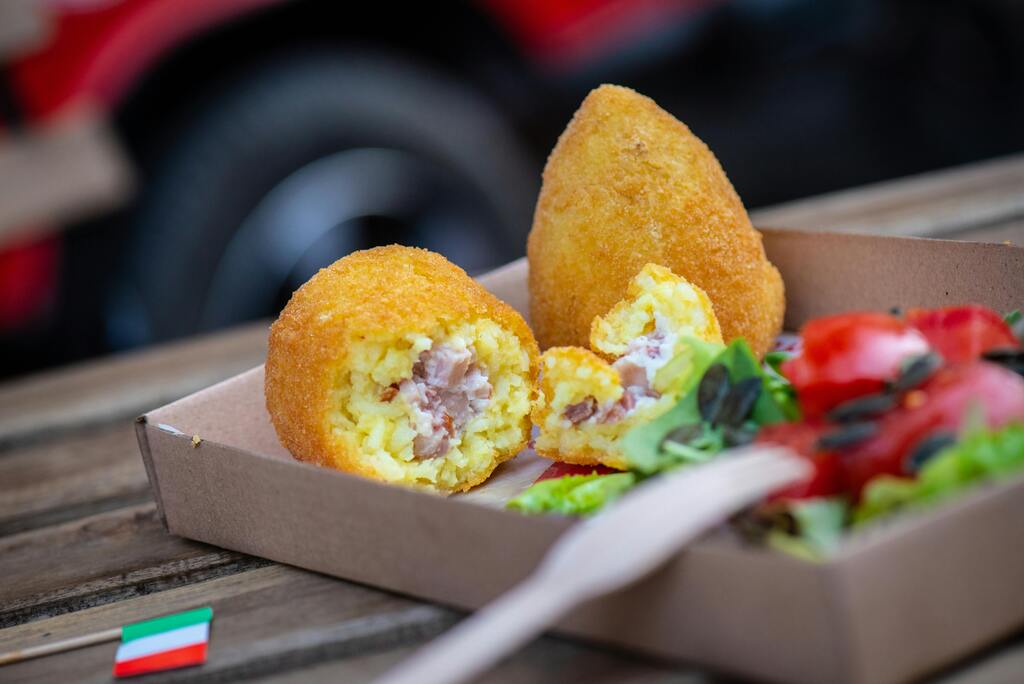 Dos arancini (bolas de arroz fritas sicilianas), uno entero y otro partido por la mitad mostrando su relleno, servidos en una caja de cartón.