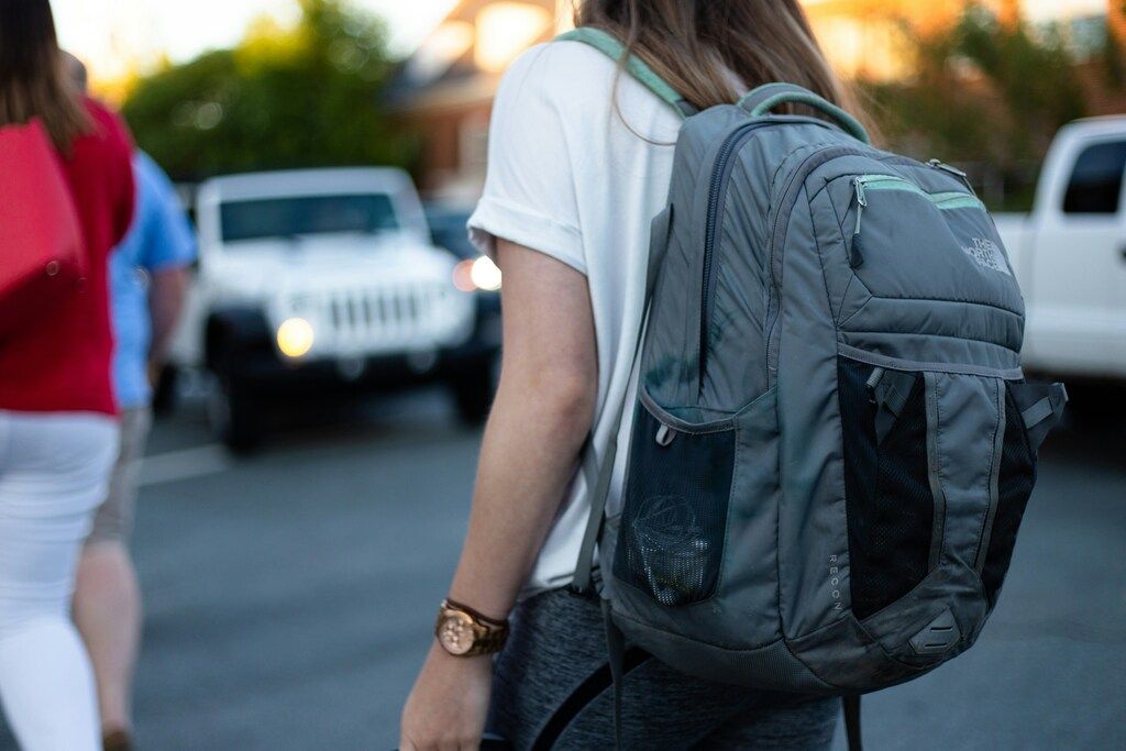 Mujer caminando con una mochila grande en la espalda, lista para un nuevo viaje, en un entorno urbano con coches al fondo.