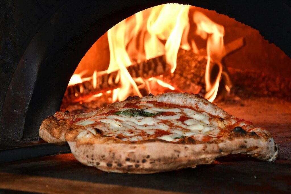 Una pizza margherita recién horneada con bordes inflados y quemados por el calor, saliendo de un horno de leña con llamas brillantes en el fondo.