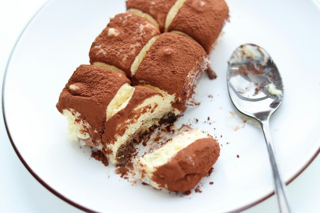 Porciones de tiramisú cubiertas con cacao en polvo en un plato blanco, con una cuchara al lado que muestra parte del postre.