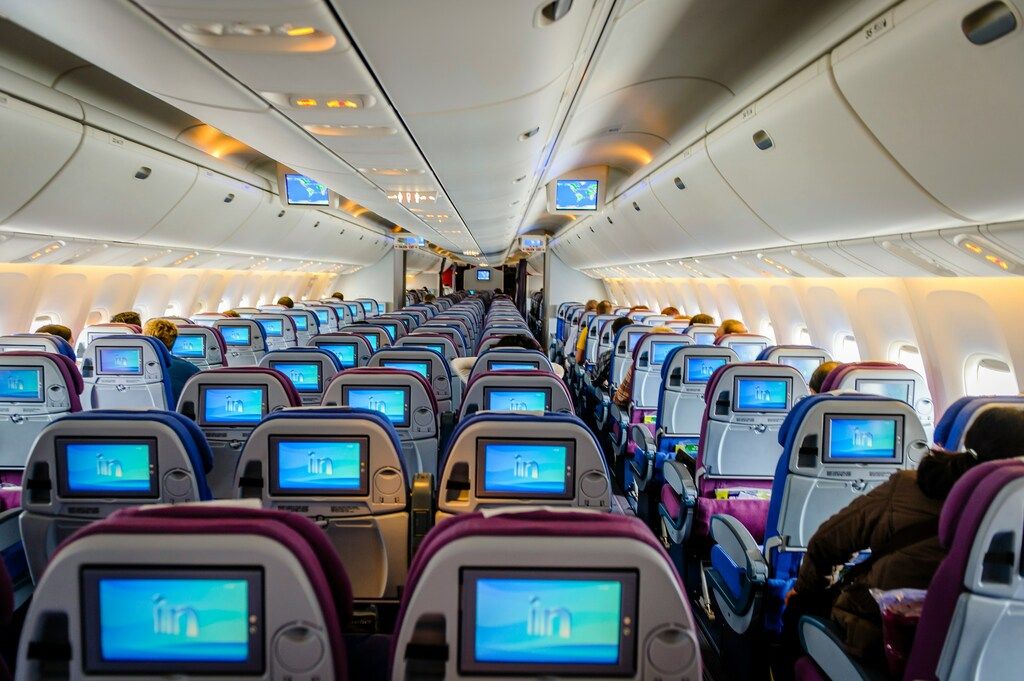 Cabina de un avión comercial con filas de asientos y pantallas individuales encendidas, vista desde la parte trasera.